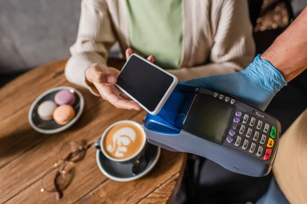 Ausgeschnittene Ansicht eines verschwommenen Kunden beim Bezahlen mit dem Smartphone in der Nähe eines afrikanisch-amerikanischen Kellners in Latexhandschuhen mit Zahlungsterminal — Stockfoto