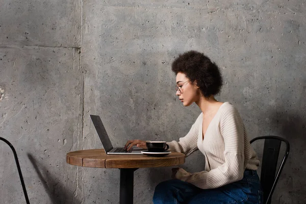 Vista lateral da mulher americana africana usando laptop perto de café no café — Fotografia de Stock