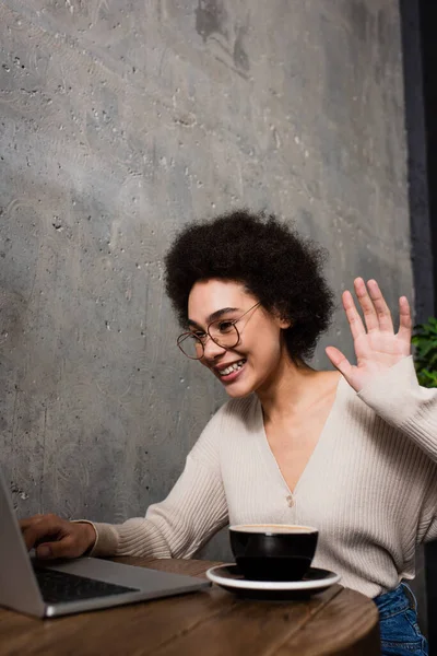Enthousiaste femme afro-américaine ayant appel vidéo sur ordinateur portable flou près du café dans le café — Photo de stock