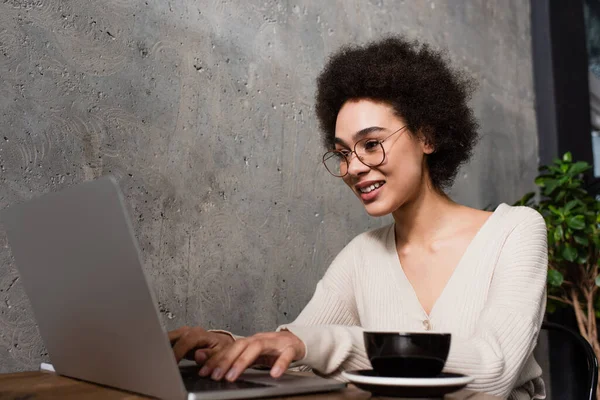 Позитивна афроамериканська телепрацівниця використовує ноутбук біля розмитої чашки в кафе. — стокове фото