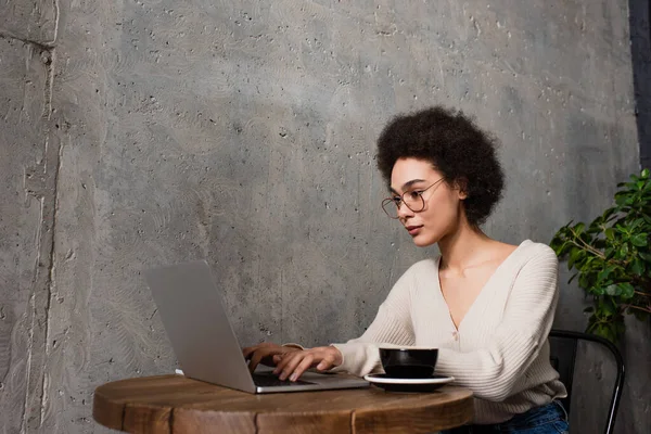 Joven afroamericano freelancer utilizando portátil cerca de taza de café en la cafetería - foto de stock