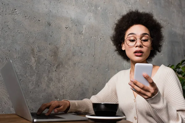 Femme afro-américaine en utilisant un ordinateur portable et smartphone près de tasse dans le café — Photo de stock