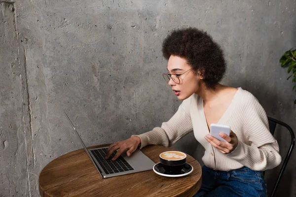 Teletrabajador afroamericano con smartphone y portátil trabajando cerca de capuchino en la cafetería - foto de stock