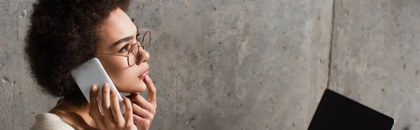 Vue latérale d'un pigiste afro-américain parlant sur smartphone près d'un ordinateur portable flou dans un café, bannière — Photo de stock