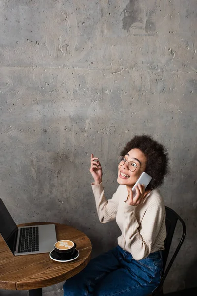 Sonriente mujer afroamericana hablando en el teléfono inteligente cerca de la computadora portátil y café en la cafetería - foto de stock