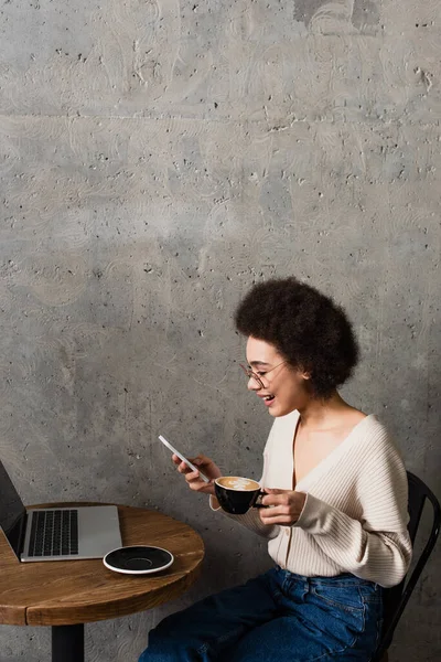 Боковой вид позитивной африканской женщины с кофе в смартфоне рядом с ноутбуком на столе в кафе — стоковое фото