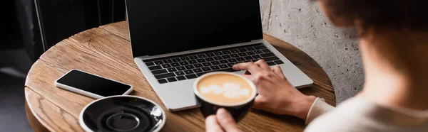 Vista recortada de la mujer afroamericana borrosa con café usando el ordenador portátil en la cafetería, pancarta - foto de stock
