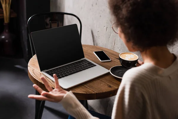 Freelancer afro-americano desfocado sentado perto de café e dispositivos no café — Fotografia de Stock