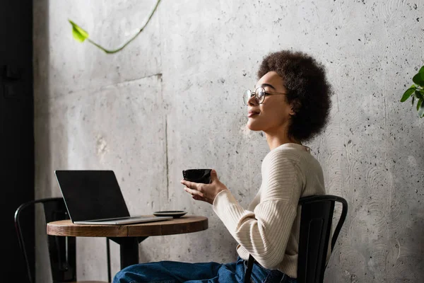 Femme afro-américaine tenant une tasse de café près d'un ordinateur portable dans un café — Photo de stock