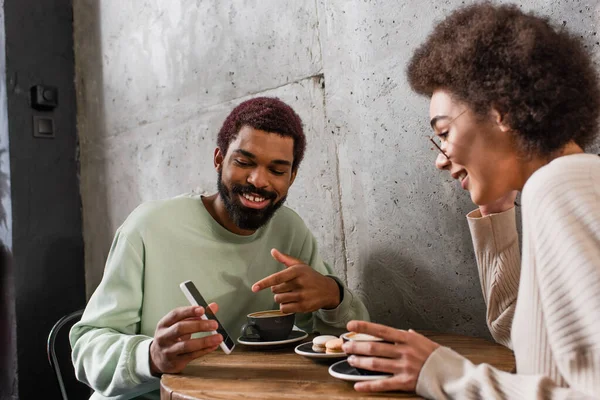 Souriant homme afro-américain pointant vers smartphone près de petite amie floue et café dans le café — Photo de stock