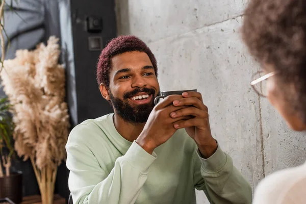 Sonriente hombre afroamericano con taza mirando a su novia en primer plano borroso en la cafetería - foto de stock