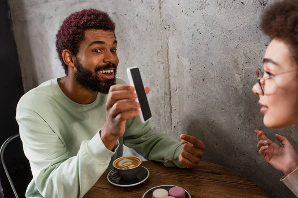 Positive african american man holding smartphone near blurred girlfriend and coffee in cafe — Stock Photo