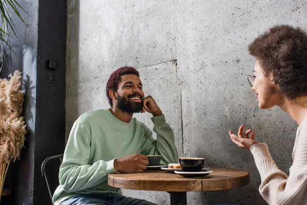 Sonriente hombre afroamericano con café mirando a su novia en la cafetería - foto de stock