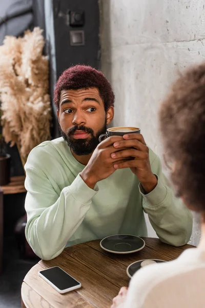 Здивований афроамериканець з кавою, що кидає на розмиту дівчину біля смартфона в кафе. — стокове фото