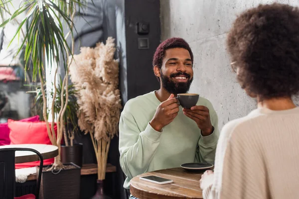 Positive african american man with coffee looking at girlfriend on blurred foreground in cafe — Stock Photo