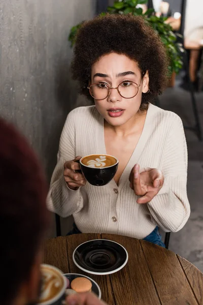 African american woman with coffee pointing at blurred boyfriend in cafe — Stock Photo