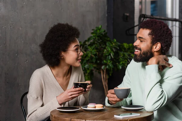 Smiling african american couple with coffee talking near macaroons in cafe — Stock Photo