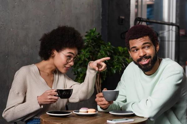 African american woman with coffee pointing with finger near smiling boyfriend in cafe — Stock Photo