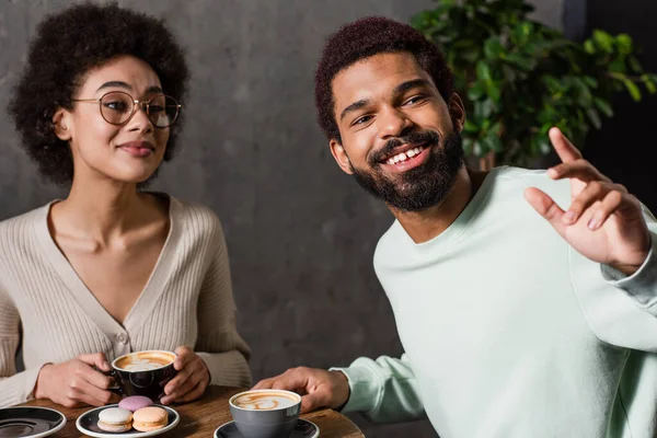 Sorrindo afro-americano homem olhando para longe perto da namorada com café e macaroons no café — Fotografia de Stock