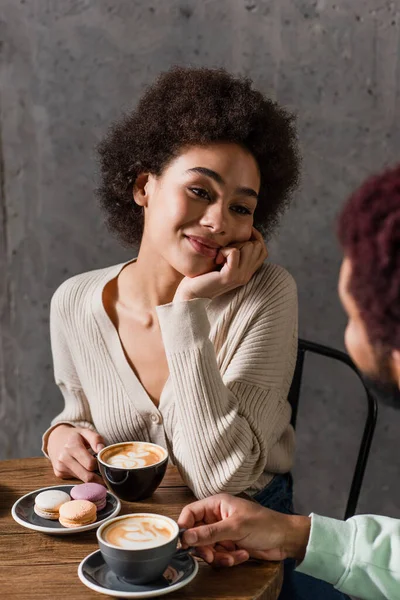 African american woman with cappuccino looking at blurred boyfriend near macaroons in cafe — Stock Photo