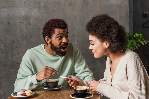 Homem americano africano animado apontando com o dedo perto da namorada com smartphone e café no café — Fotografia de Stock
