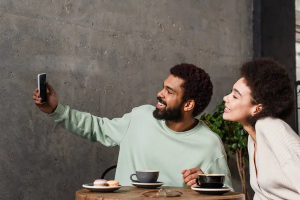 Vista lateral de la sonriente pareja afroamericana tomando selfie cerca de café y macarrones en la cafetería - foto de stock