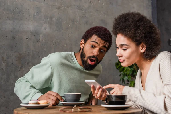 Amazed uomo africano americano seduto vicino fidanzata con smartphone e caffè in caffè — Foto stock