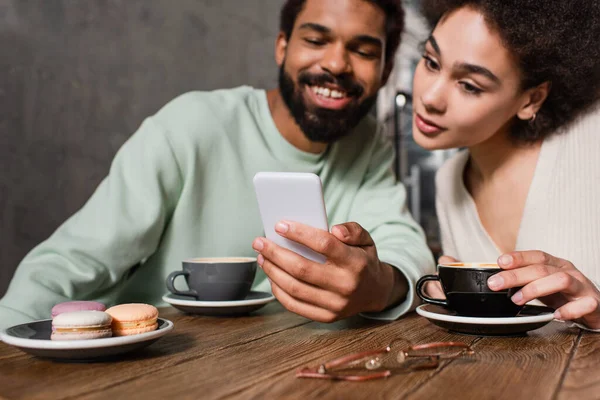 Smartphone en main de sourire homme afro-américain près de petite amie avec du café et des macarons dans le café — Photo de stock