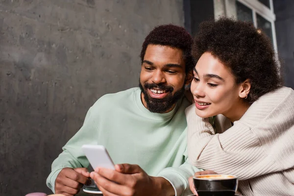 Positive african american couple using blurred smartphone near coffee in cafe — Stock Photo