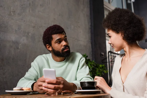 Hombre afroamericano con teléfono inteligente mirando a su novia cerca del café y macarrones borrosos en la cafetería - foto de stock