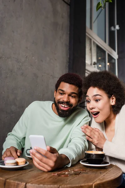 Aufgeregtes afrikanisch-amerikanisches Paar nutzt Smartphone in der Nähe von Makronen und Kaffee im Café — Stockfoto