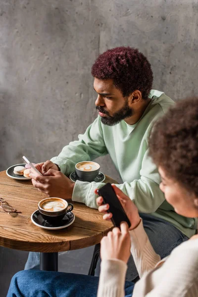 Uomo afroamericano utilizzando smartphone vicino tazze di caffè e fidanzata offuscata — Foto stock