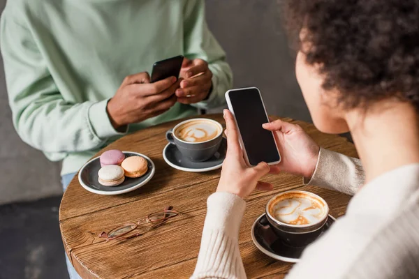 Smartphone in hands of african american woman near cappuccino and blurred boyfriend in cafe — Stock Photo