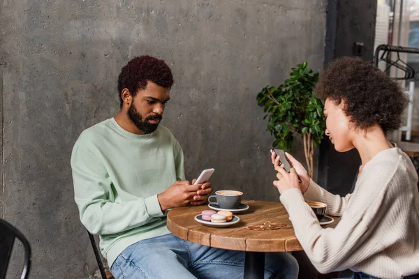 Junges afrikanisch-amerikanisches Paar nutzt Smartphones in der Nähe von Makronen und Kaffee im Café — Stockfoto