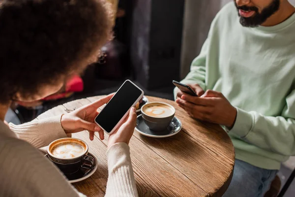 Smartphone avec écran vierge dans les mains d'une femme afro-américaine assise près du petit ami et du café dans un café — Photo de stock