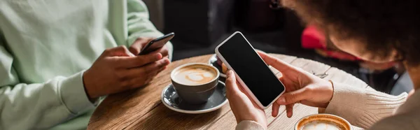 Smartphone nelle mani di donna afro-americana vicino fidanzato offuscata e caffè in caffè, banner — Foto stock