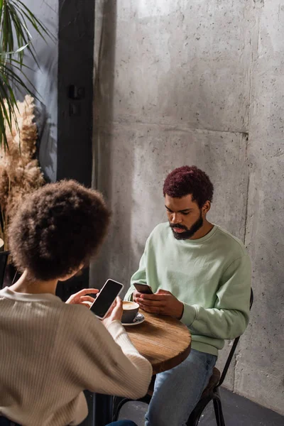 African american man using smartphone near cup of coffee and girlfriend in cafe — Stock Photo