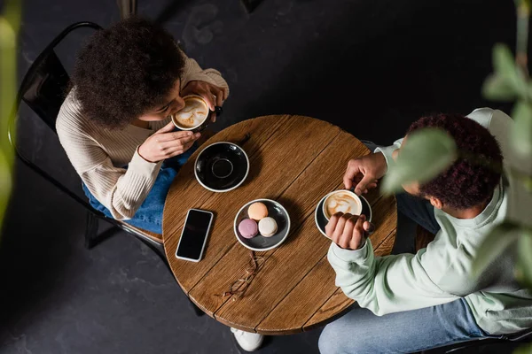 Vista aerea della giovane coppia afro-americana che beve caffè vicino ai macaron e allo smartphone nel caffè — Foto stock