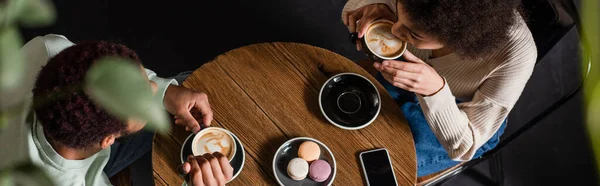 Vista aerea della donna afroamericana che beve cappuccino vicino al fidanzato, smartphone e macaron in caffè, banner — Foto stock