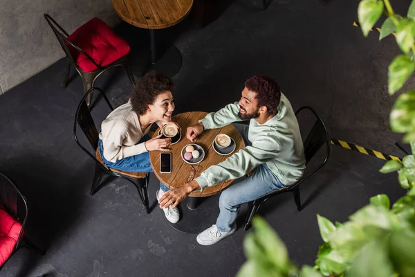 Vue aérienne de heureux couple afro-américain parlant près de café, macarons et smartphone dans le café — Photo de stock