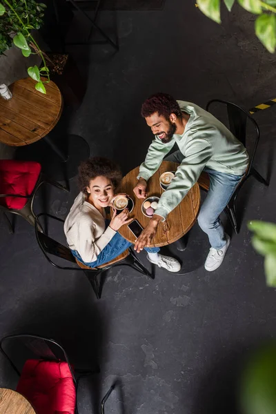 Vista aerea dell'uomo afro-americano sorridente che agita la mano vicino alla ragazza con il caffè nel caffè — Foto stock