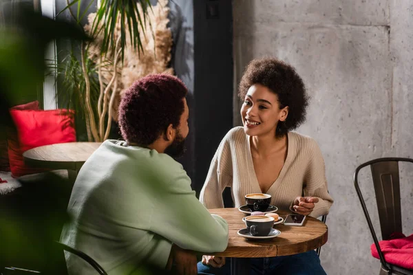 Mujer sonriente hablando con novio afroamericano cerca de café y teléfono inteligente - foto de stock