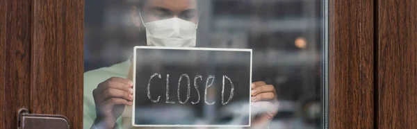 African american barista in medical mask holding chalkboard with closed lettering near door of cafe, banner — Stock Photo