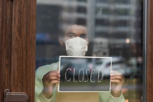 Barista afroamericano en máscara médica sosteniendo pizarra con letras cerradas en la cafetería - foto de stock