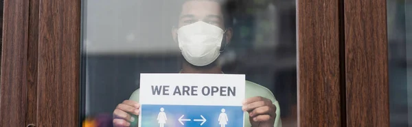 Barista afroamericano en máscara médica sosteniendo letrero con estamos letras abiertas cerca de la puerta de la cafetería, pancarta - foto de stock