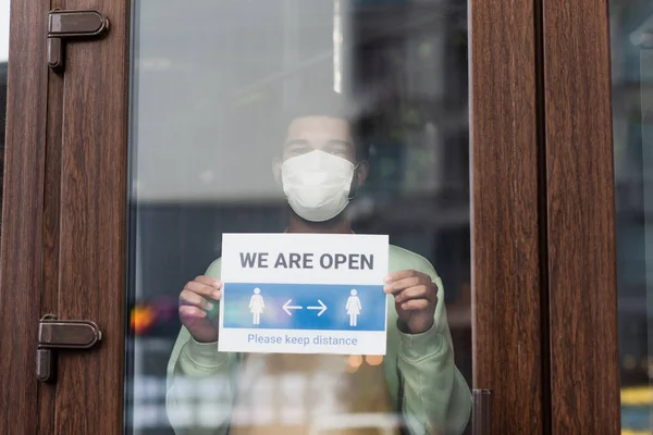 African american barista in medical mask holding signboard with we are open lettering near door of cafe — Stock Photo