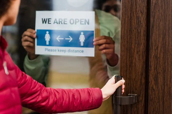 Vista recortada de la puerta de apertura del cliente cerca de barista afroamericano con estamos letras abiertas en el letrero en la cafetería - foto de stock