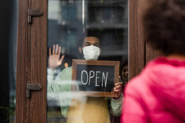 African american barista in medical mask holding signboard with open lettering in cafe near blurred client — Stock Photo