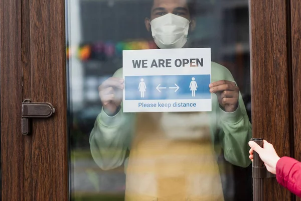 Barista afroamericano in maschera medica che tiene cartello con siamo aperti lettering vicino al cliente in caffè — Foto stock