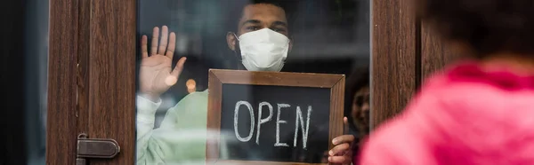 African american barista in medical mask holding chalkboard with open lettering and waving hand near blurred client in cafe, banner — Stock Photo